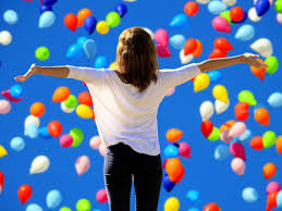 Girl facing hundreds of floating balloons, arms spread with joy